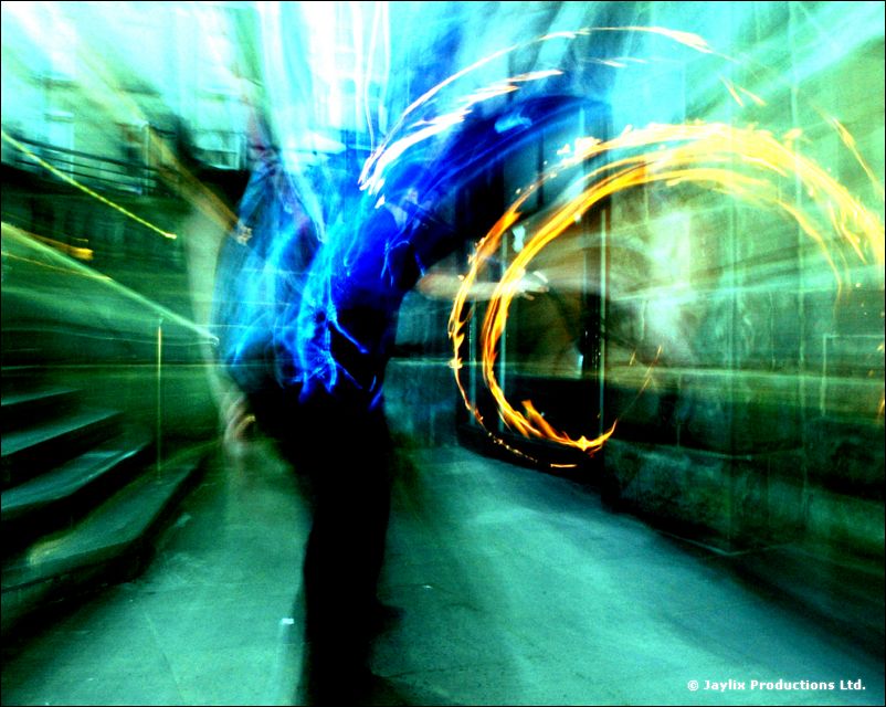 A blurred figure and a curved trail of fire in a green and blue lit alley