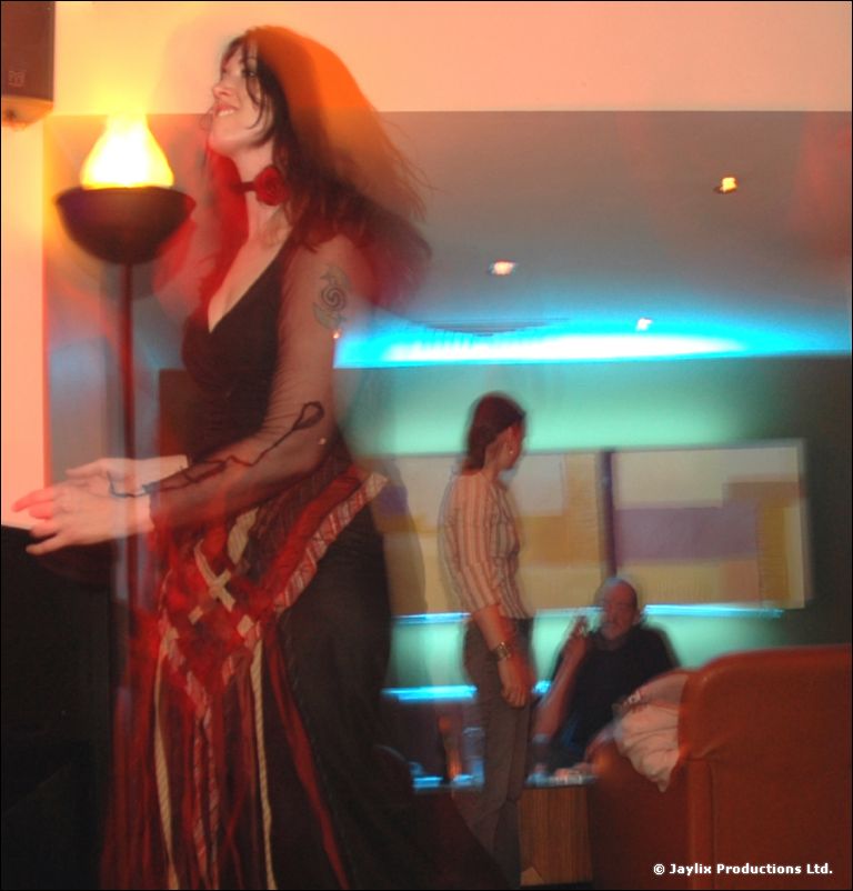 A person in a long black and red dress stands near a flame lamp in a lounge, blurred with motion