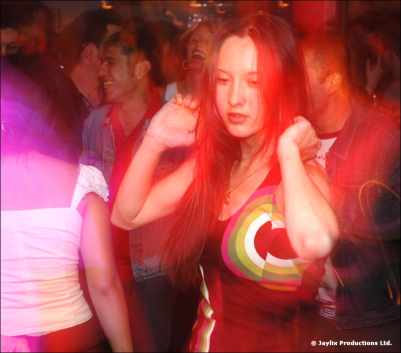 A woman with long hair and a colourful target-design top dances in a crowd, lit red