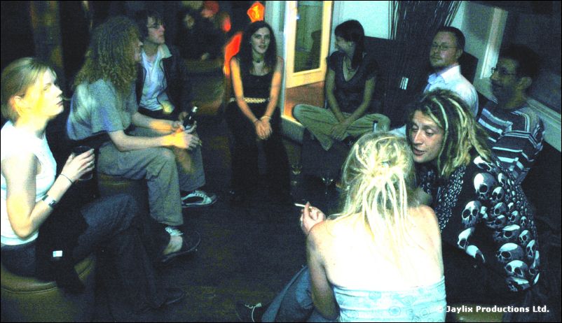 A group sits in a circle on the floor and stools in a dim room, some smoking