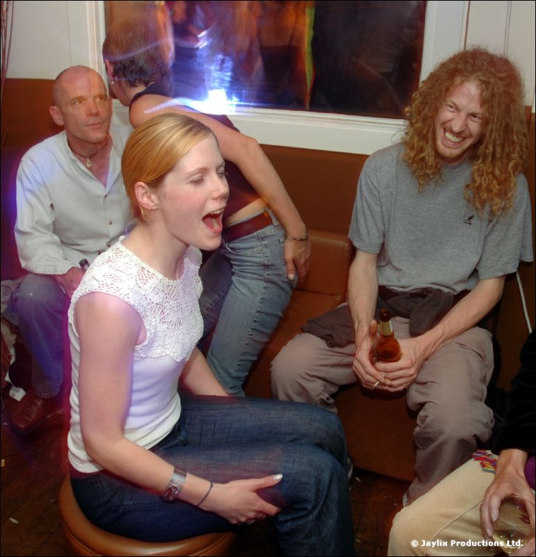 Five people sit on a bench seat, a woman in white lace top singing, a curly-haired man laughing