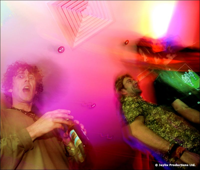 Two blurred men, one in a green patterned shirt, dance under pink and magenta light