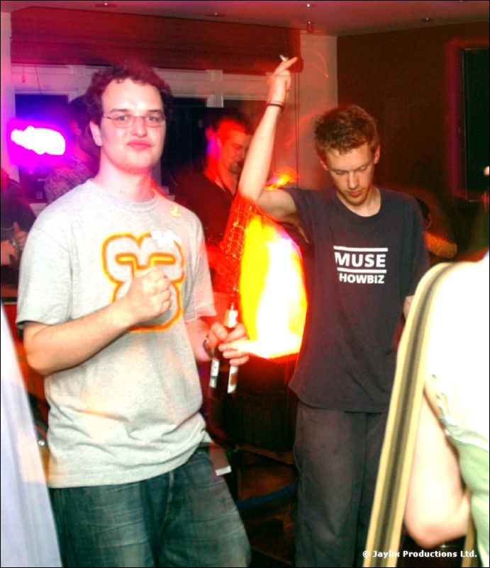 Two men stand with drinks in a bar, one in a grey t-shirt and one in a black Muse t-shirt