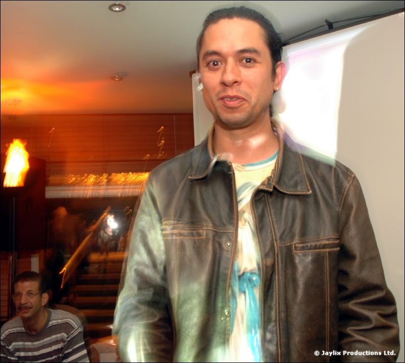 A man in a brown leather jacket stands in a bar near a projector screen