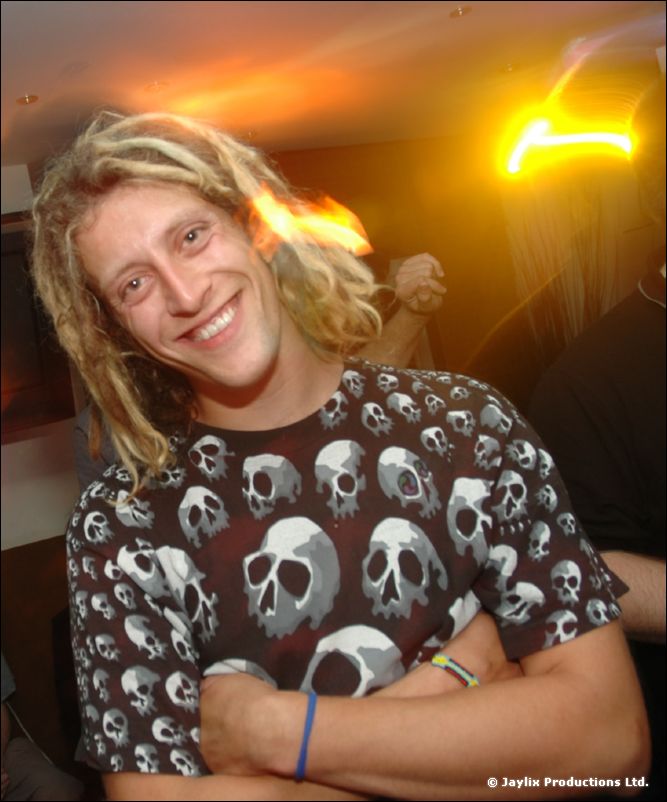 A smiling person with long blond dreadlocks in a skull-print t-shirt, arms crossed