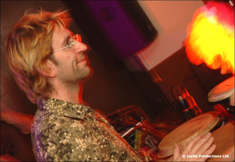 A man with glasses and a patterned shirt plays congas, with a flame blurred behind him