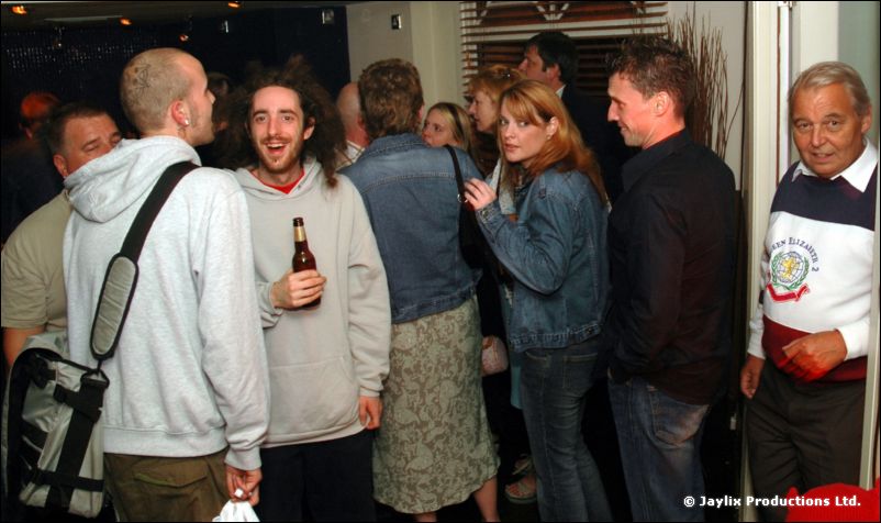A standing crowd in a bar, a man holding a beer bottle and an older man in a striped jumper