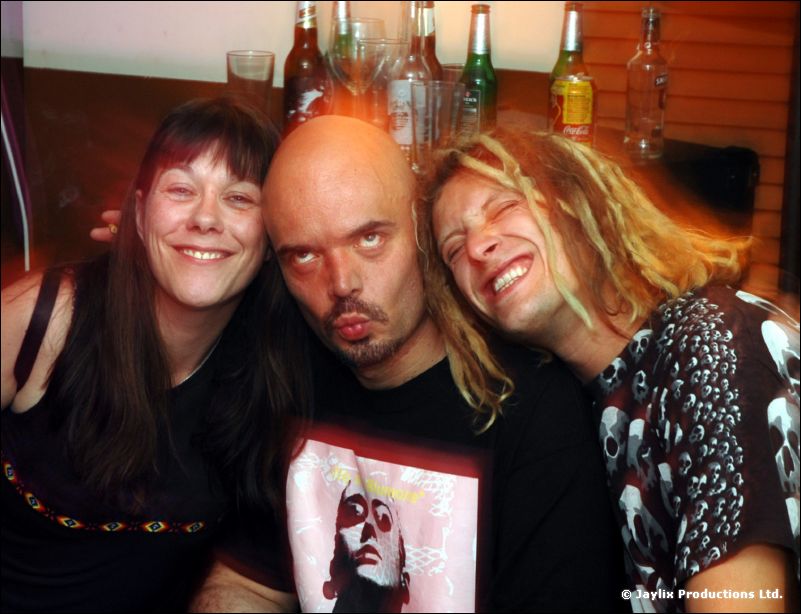 Three people pose close together in front of a bar, the middle man pulling a face
