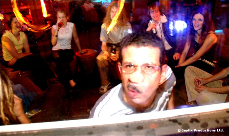 A man with glasses and a moustache leans forward at a table, people seated behind him