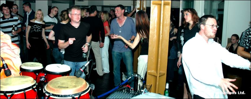 People stand around bongo drums in a bar, with a man in a white shirt gesturing at right