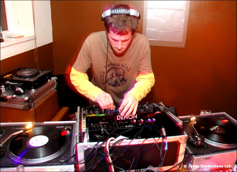 A DJ in headphones and a beige long-sleeved top mixes at turntables and a Denon mixer
