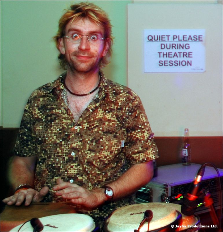 A man with wide eyes plays congas beside a sign reading Quiet Please During Theatre Session