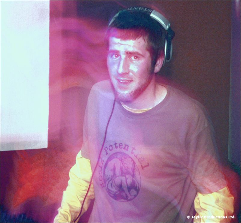 A DJ in headphones and a purple t-shirt looks at the camera, lit in pink and violet