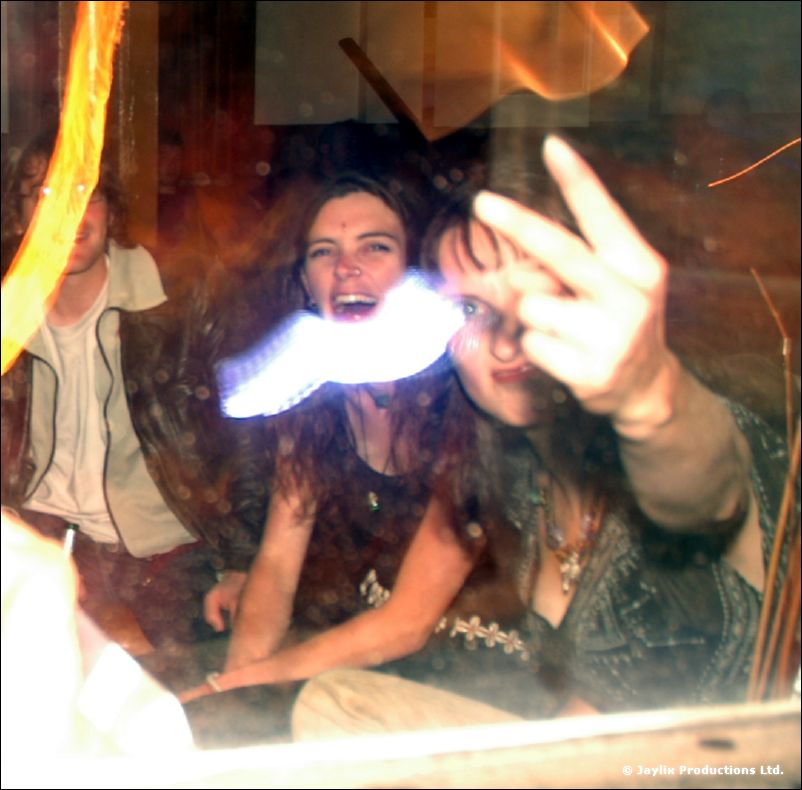 Two women smile through a glass pane as one makes a rude hand gesture, with a glowing blur in front