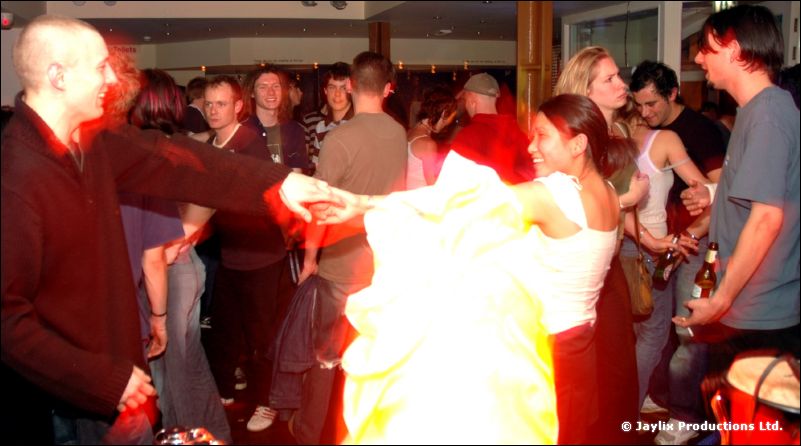 A man and a woman dance holding hands in a crowded room, a bright white blur between them