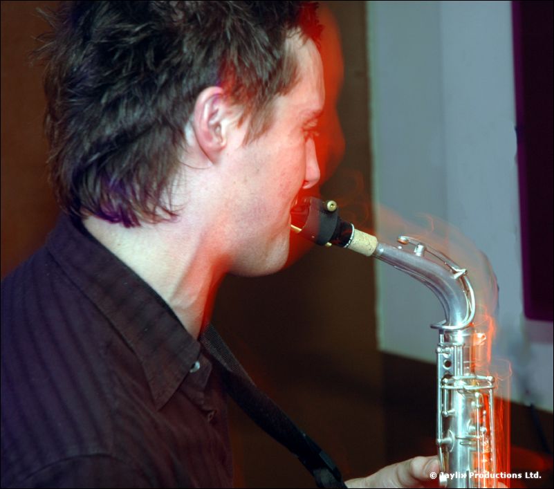 A man in a dark shirt plays a silver saxophone, seen in profile