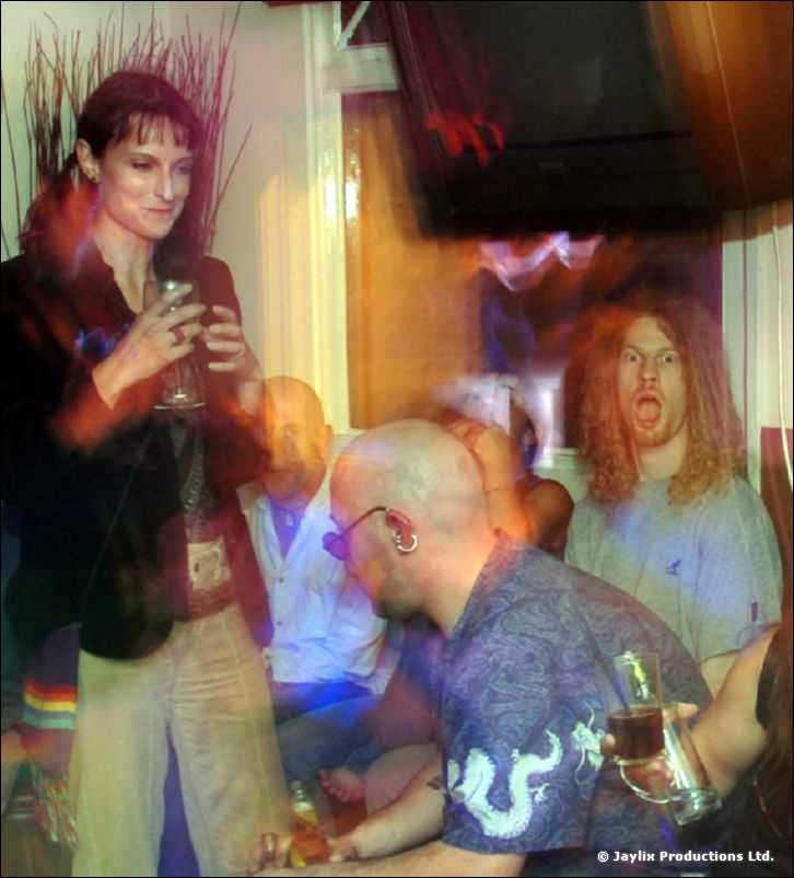 People stand and sit in a bar, a bald man in a blue dragon shirt and a curly-haired man behind him