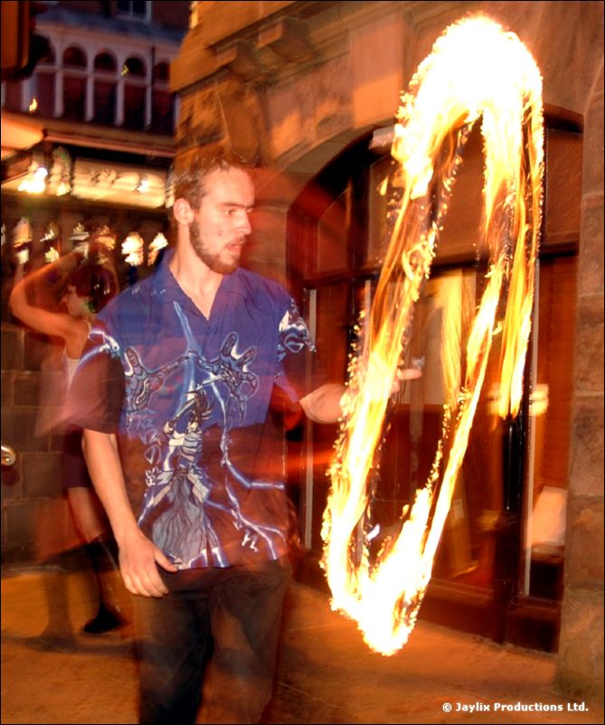 A man in a blue patterned shirt stands by a large ring of fire on a street at night
