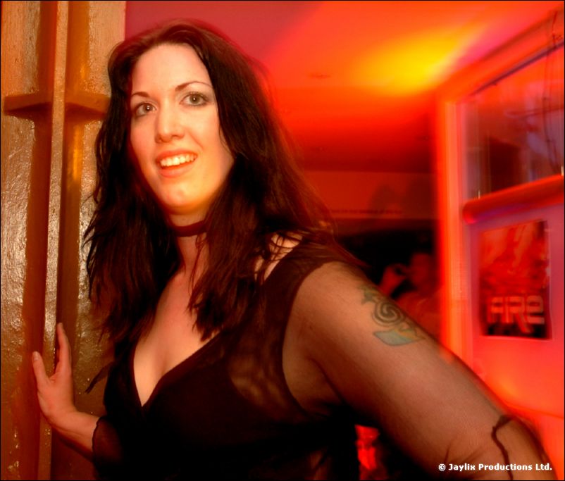 A smiling woman with long dark hair and a tattooed arm stands in an orange-lit doorway, a FIRE poster behind her