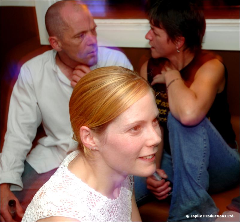A woman in a white lace top in front, with a bald man and a woman behind her on a bench
