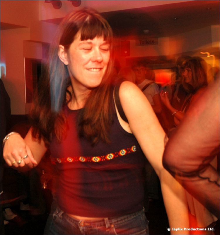 A smiling woman with long hair and a dark top dances in a red-lit bar