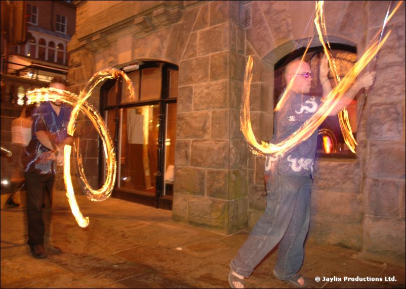 Two people spin fire outside a stone building at night, one in a blue shirt and one in jeans