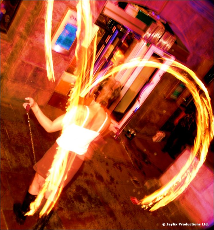 A person in a white top spins loops of fire in a tilted shot of a street at night