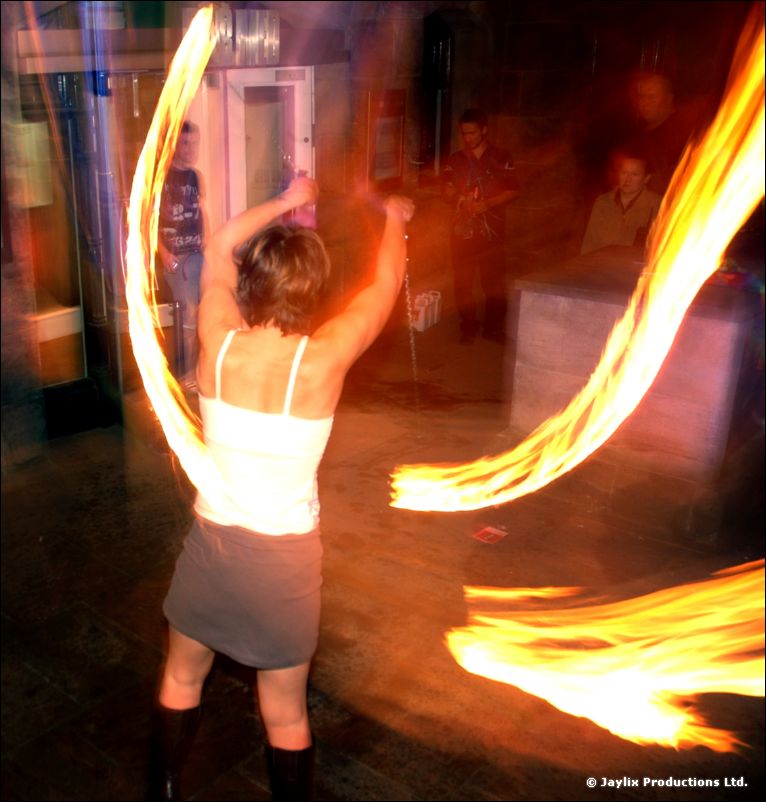 A person in a white top and grey skirt spins fire in a dark bar, arms raised