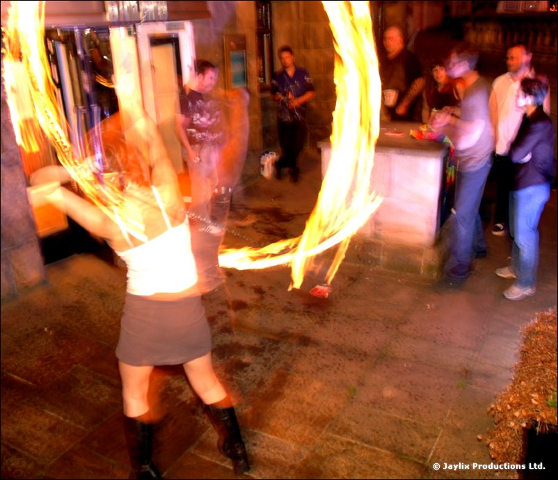 A person in a white top and grey skirt spins fire outside a bar, with people standing behind