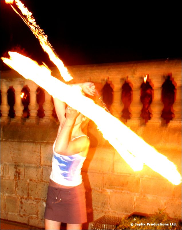 A person in a white top and grey skirt swings a flaming staff in front of a stone balustrade at night