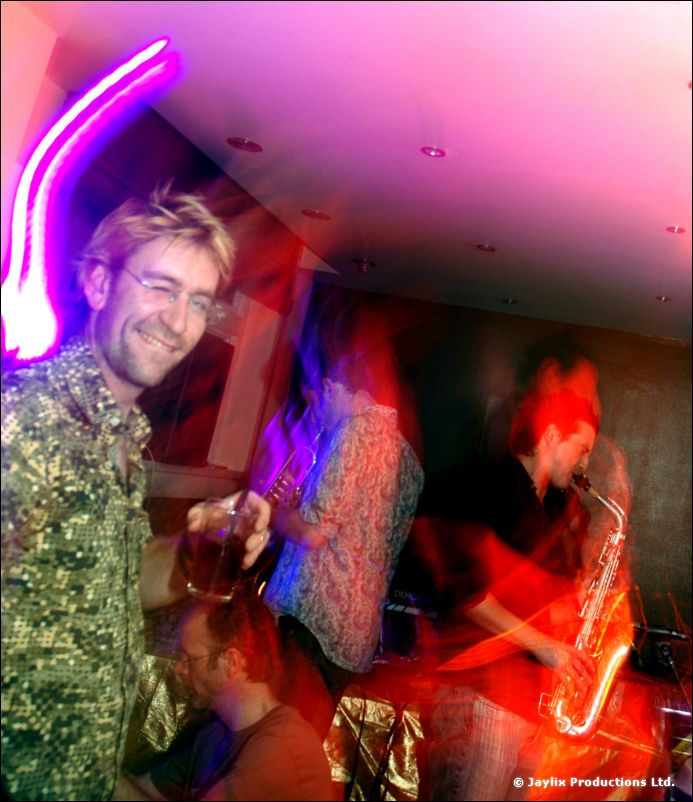 A smiling man in a green patterned shirt with a band playing saxophone behind him, pink light above