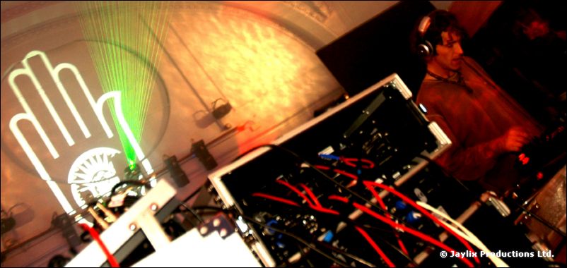 A DJ wearing headphones behind a mixer with red cables, beside a projected white hand logo and green laser