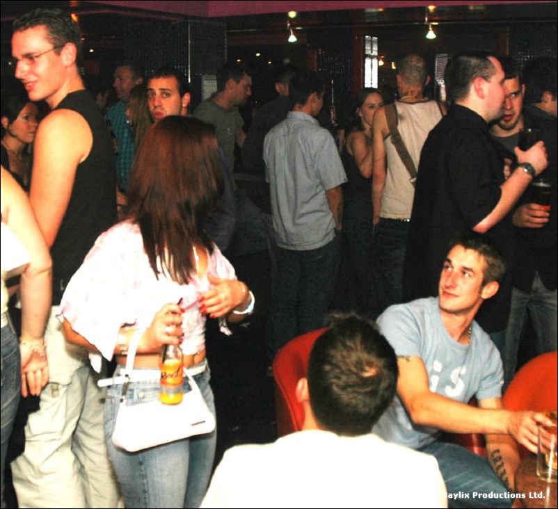 A crowded bar with people standing and chatting, one person seated in a grey t-shirt looking up
