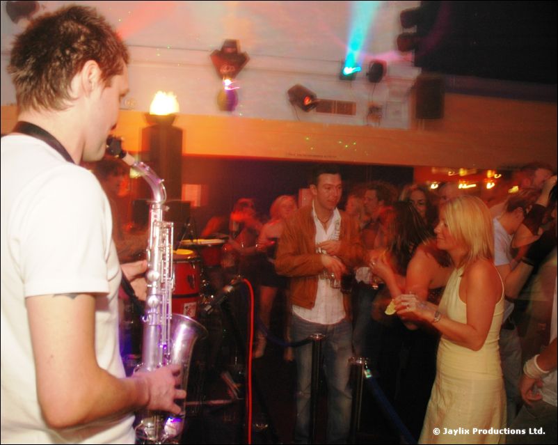 A person in a white t-shirt plays a saxophone with a crowd dancing behind