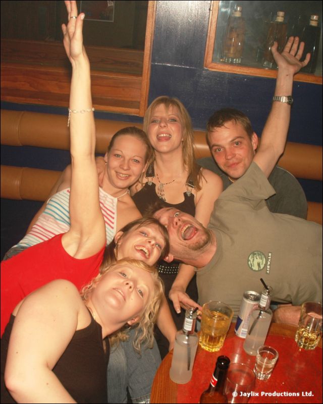 A group of people crowded round a table with drinks, one raising an arm and laughing