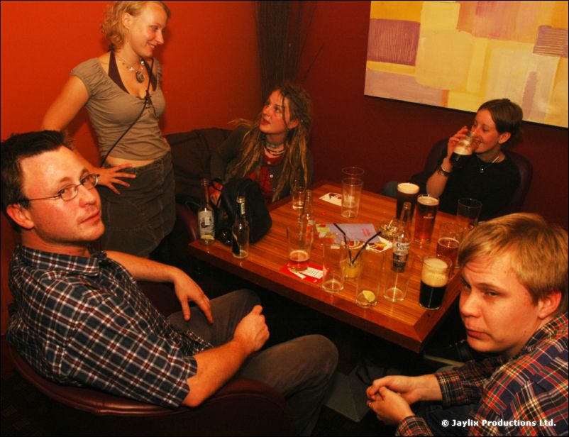 Five people around a wooden table with pints in a red-walled bar, two of them looking at the camera