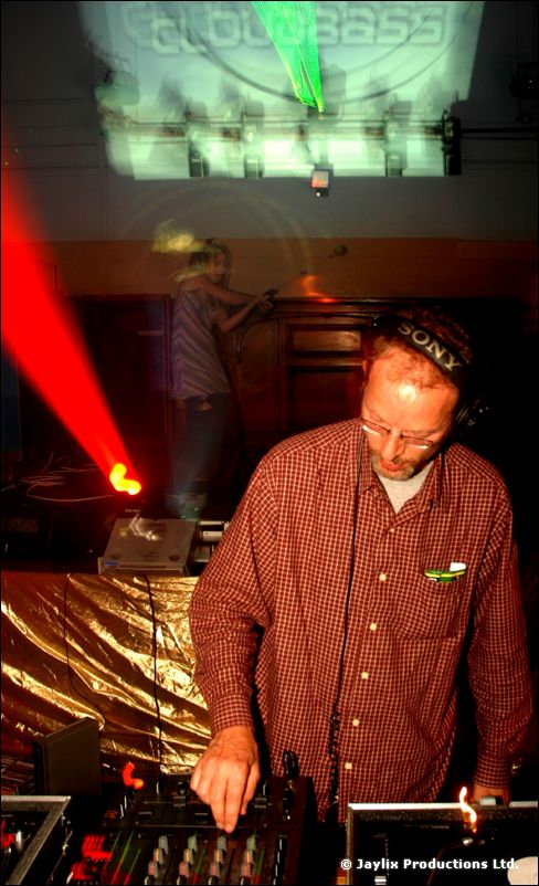 A DJ in a red checked shirt and headphones at the decks, with a laser beam and projected text behind