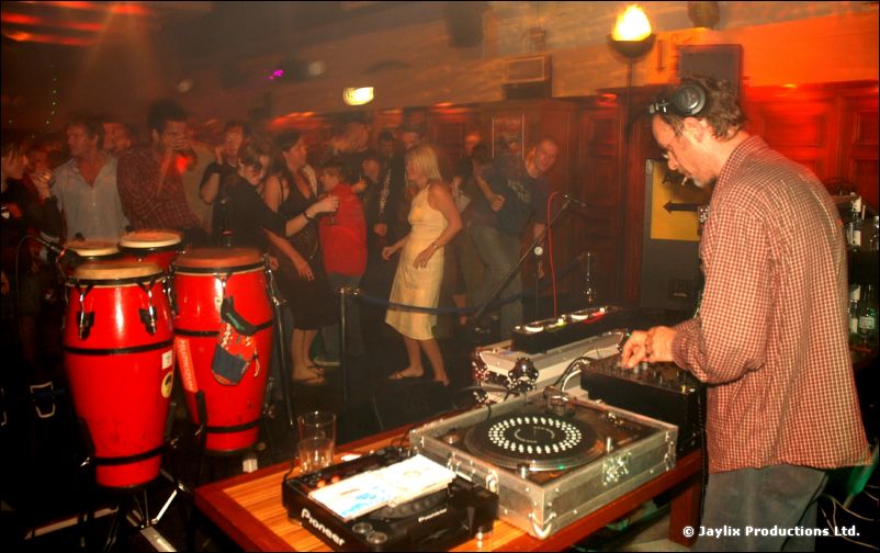 A DJ in a striped shirt and headphones at the decks, with conga drums and a crowd behind