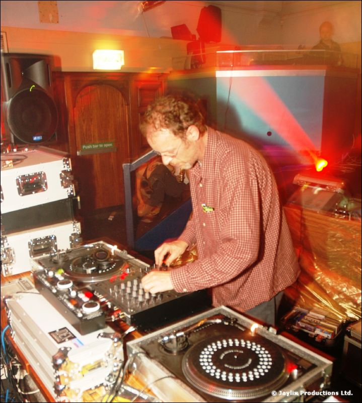 A DJ in a red checked shirt mixing at turntables, with a red laser beam and speaker behind