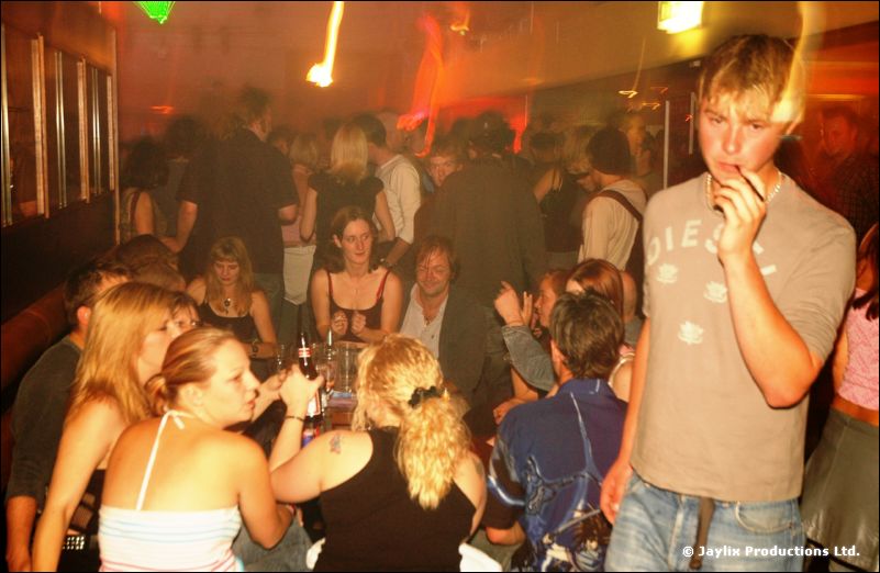 A young person in a grey t-shirt stands in front of a crowd seated and standing in a busy bar