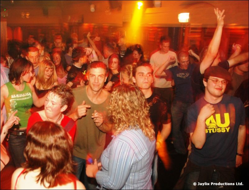 Dancers in a crowd, one raising an arm, under orange stage lighting