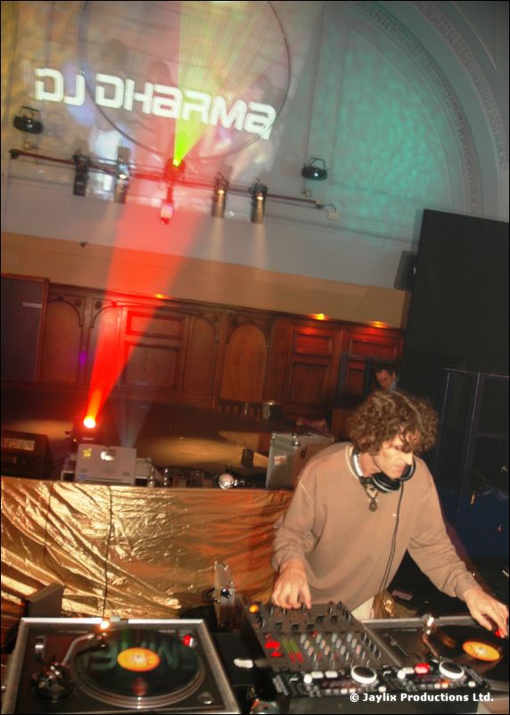 A DJ with curly hair at turntables, with "DJ Dharma" projected on the wall behind