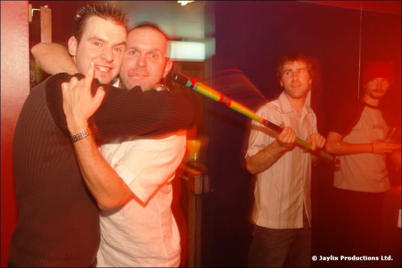 Two men embracing in red light, with two other people behind and a striped tube held between them