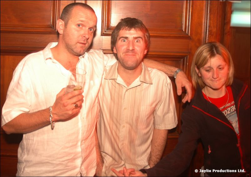 Two men in light shirts and a woman in a dark jacket pose together against a wood panelled wall