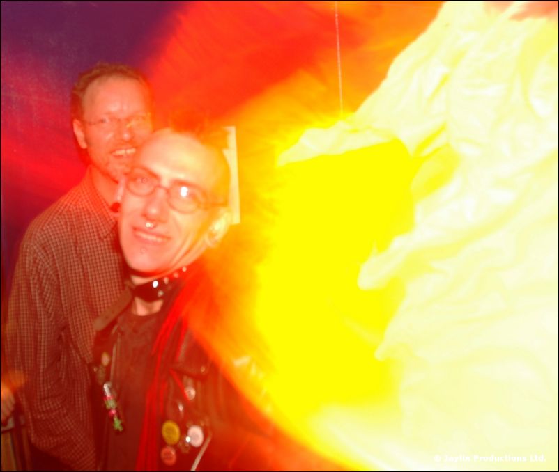Two men with glasses, one wearing badges on a dark jacket, blurred by a bright yellow flare