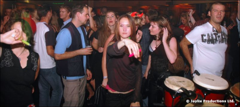 People standing in a crowded room, one pointing at the camera, with bongo drums in front