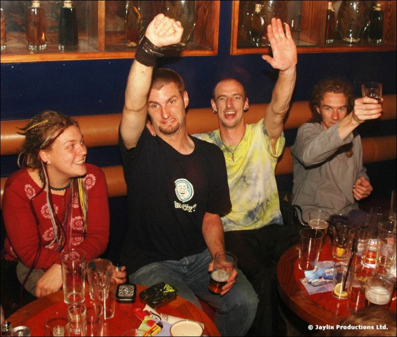 Four people at a table with drinks, two raising their arms, in front of a blue wall