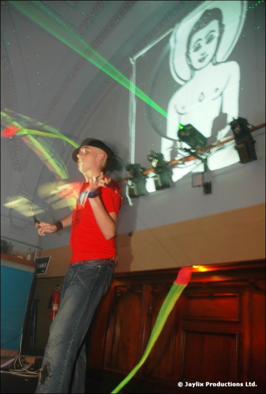 A person in a red top swings a glowing prop in front of a projected cartoon face and green laser lines