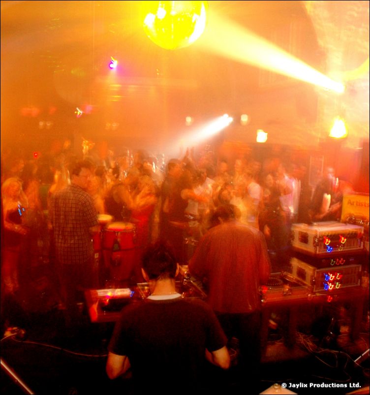 A large crowd dances in an orange haze under a mirror ball, with a DJ seen from behind