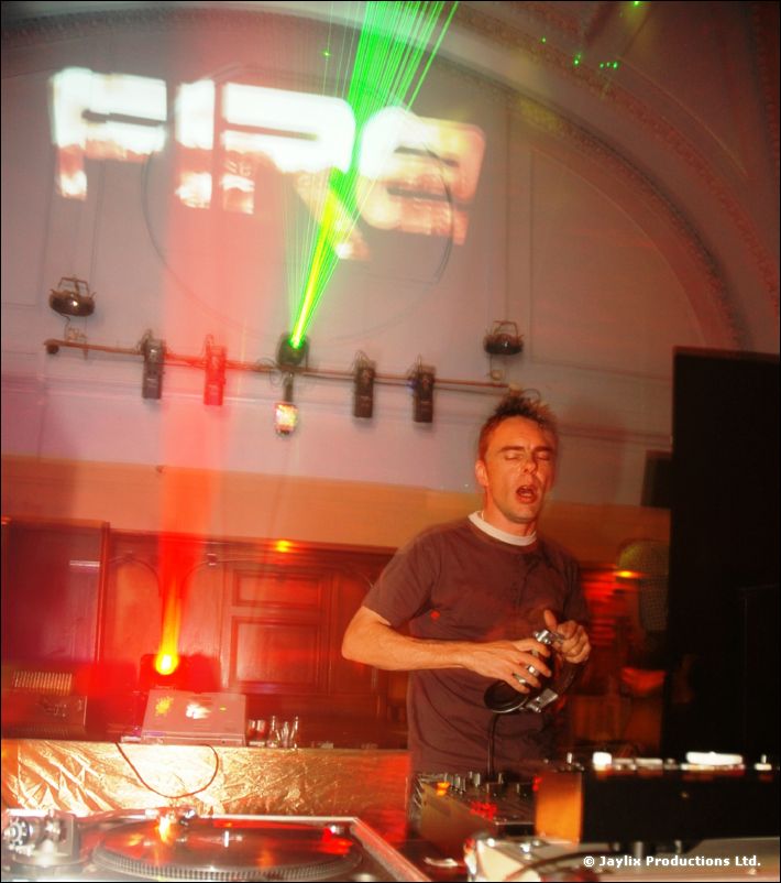 A DJ in a dark t-shirt with eyes closed at the decks, with a projected "FIRE" logo and green laser behind